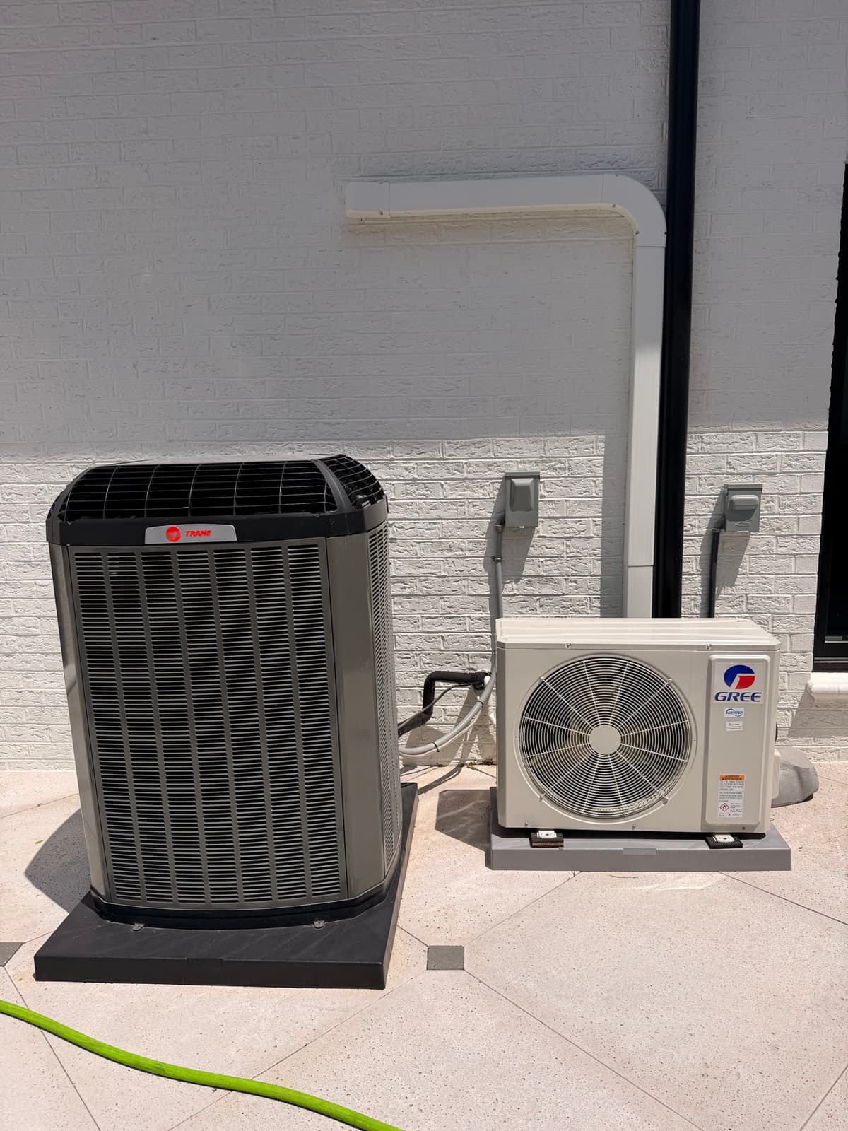 Trane central-air condenser and Gree ductless outdoor unit beside a brick wall